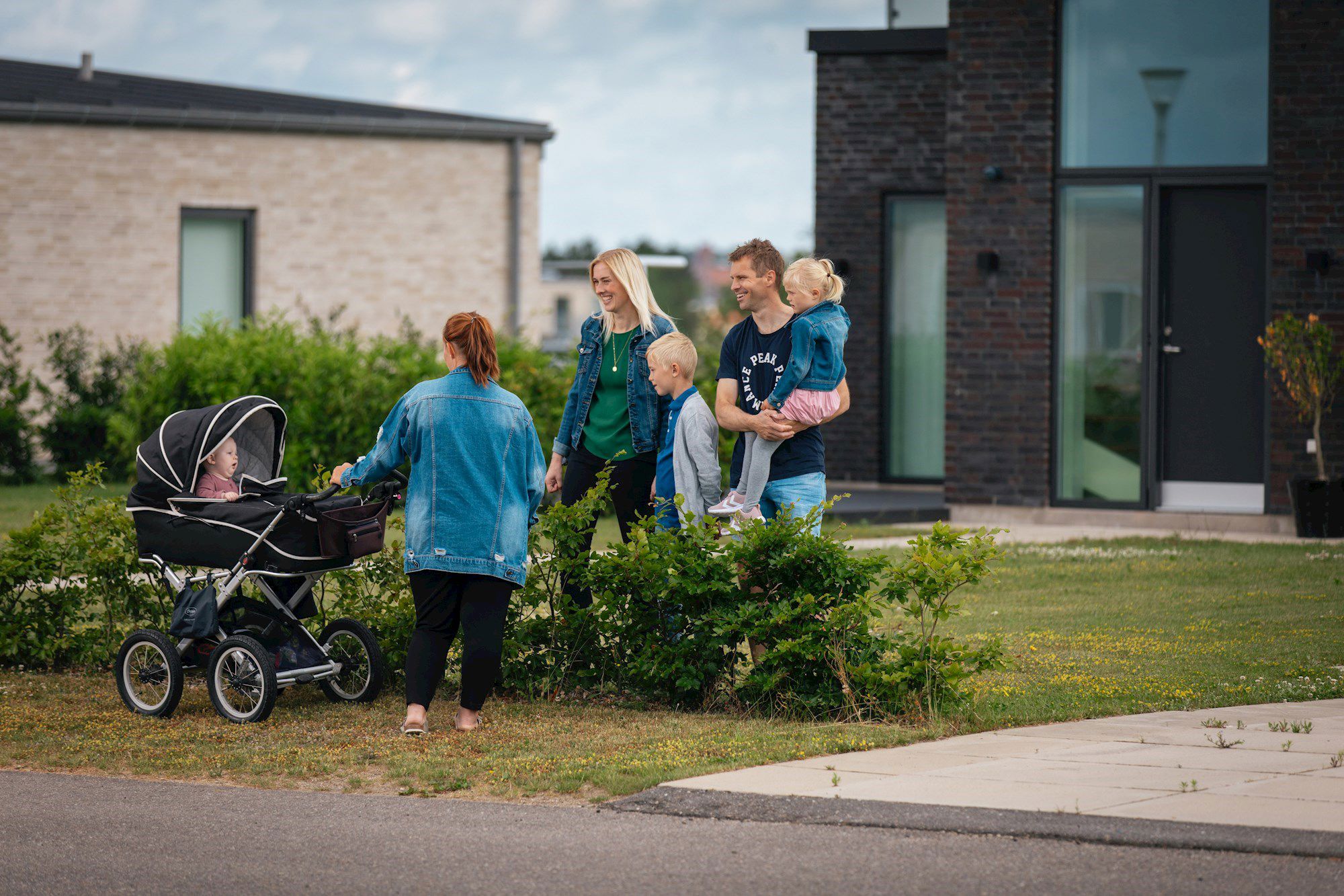 Glad forældrepar med 2 mindre børn og mor med barn i barnevogn set foran to nyere huse