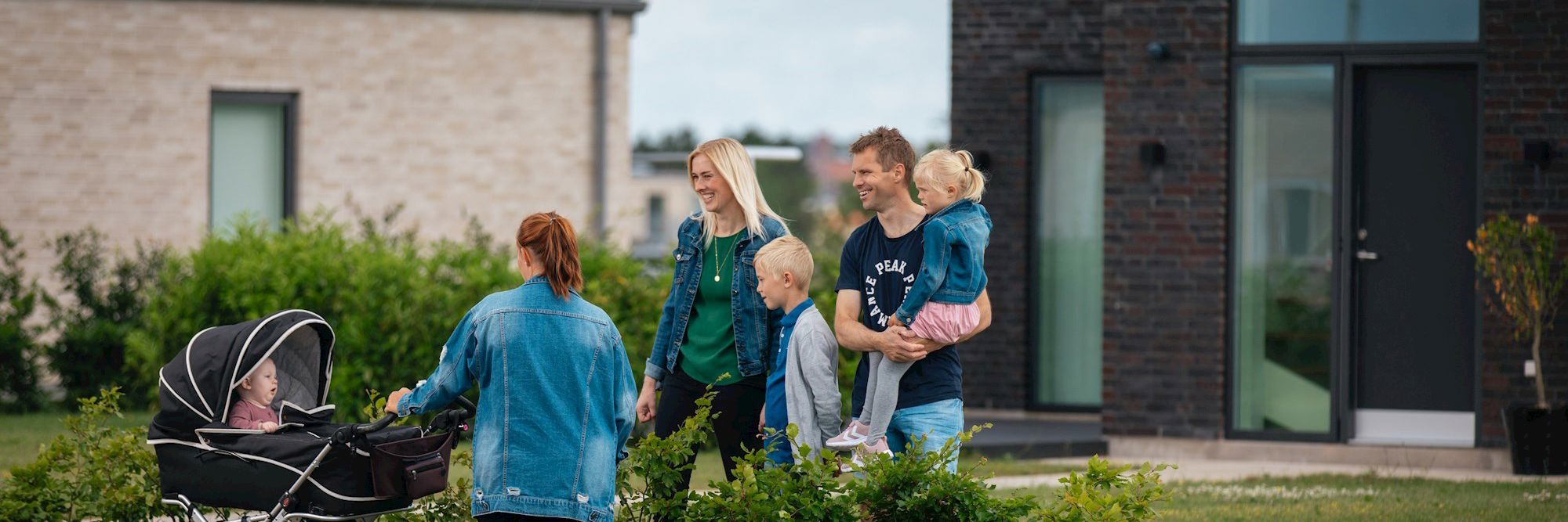Glad forældrepar med 2 mindre børn og mor med barn i barnevogn set foran to nyere huse