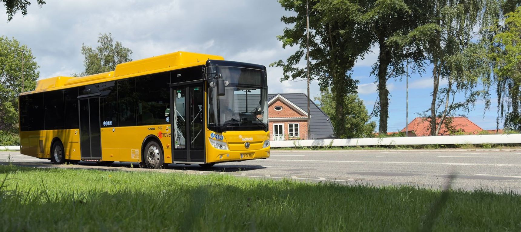 Gul bybus på en vej. Huse og træer i baggrunden.