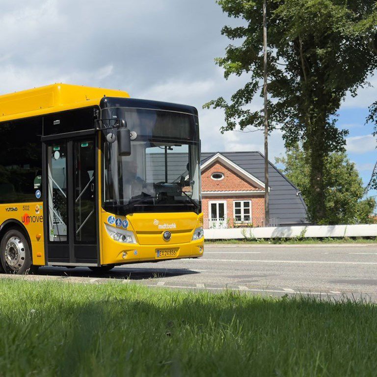 Gul bybus på en vej. Huse og træer i baggrunden.