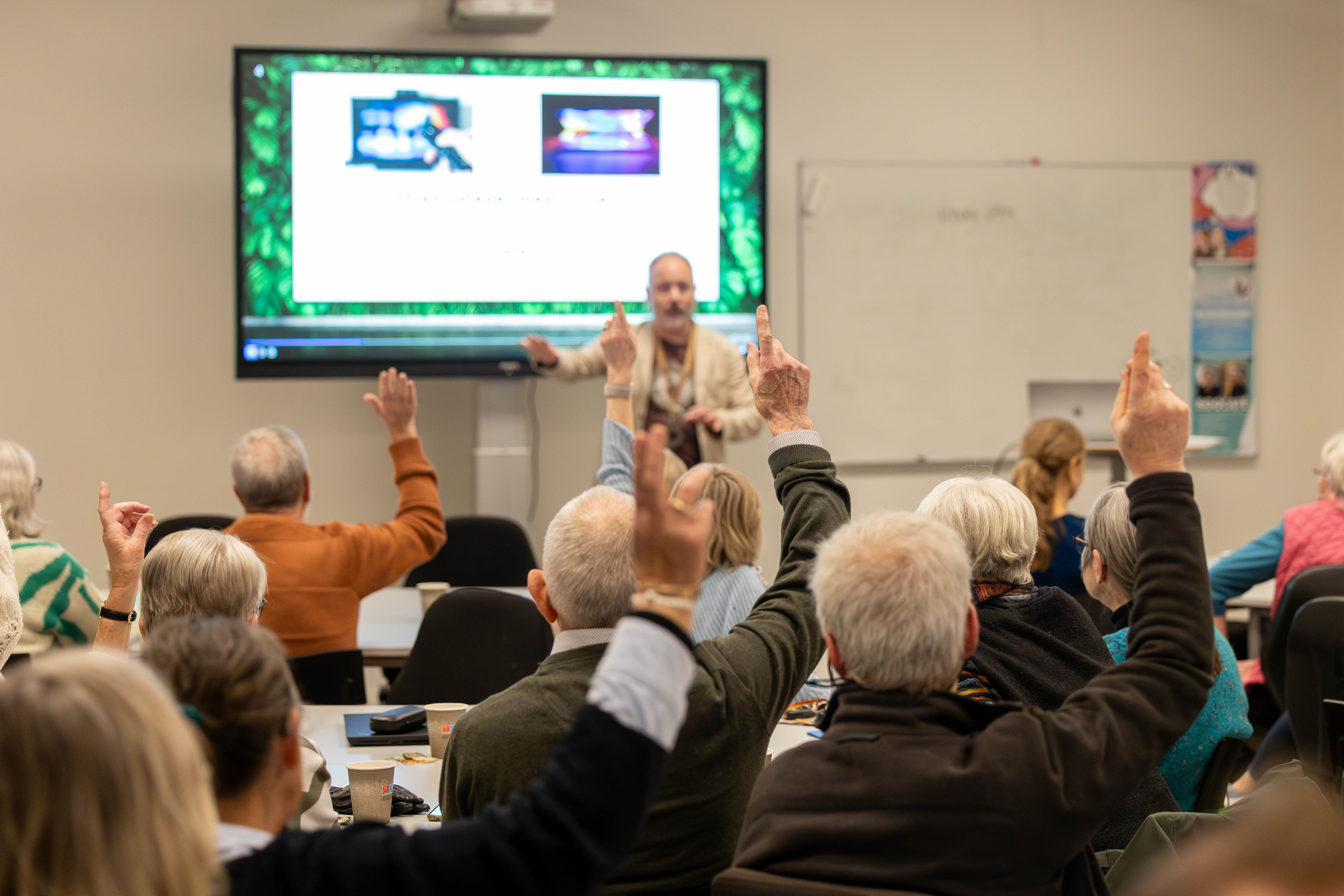 Møde. En mand står med fronten mod kamera og deltagere sidder med ryggen til og rækker hånden op