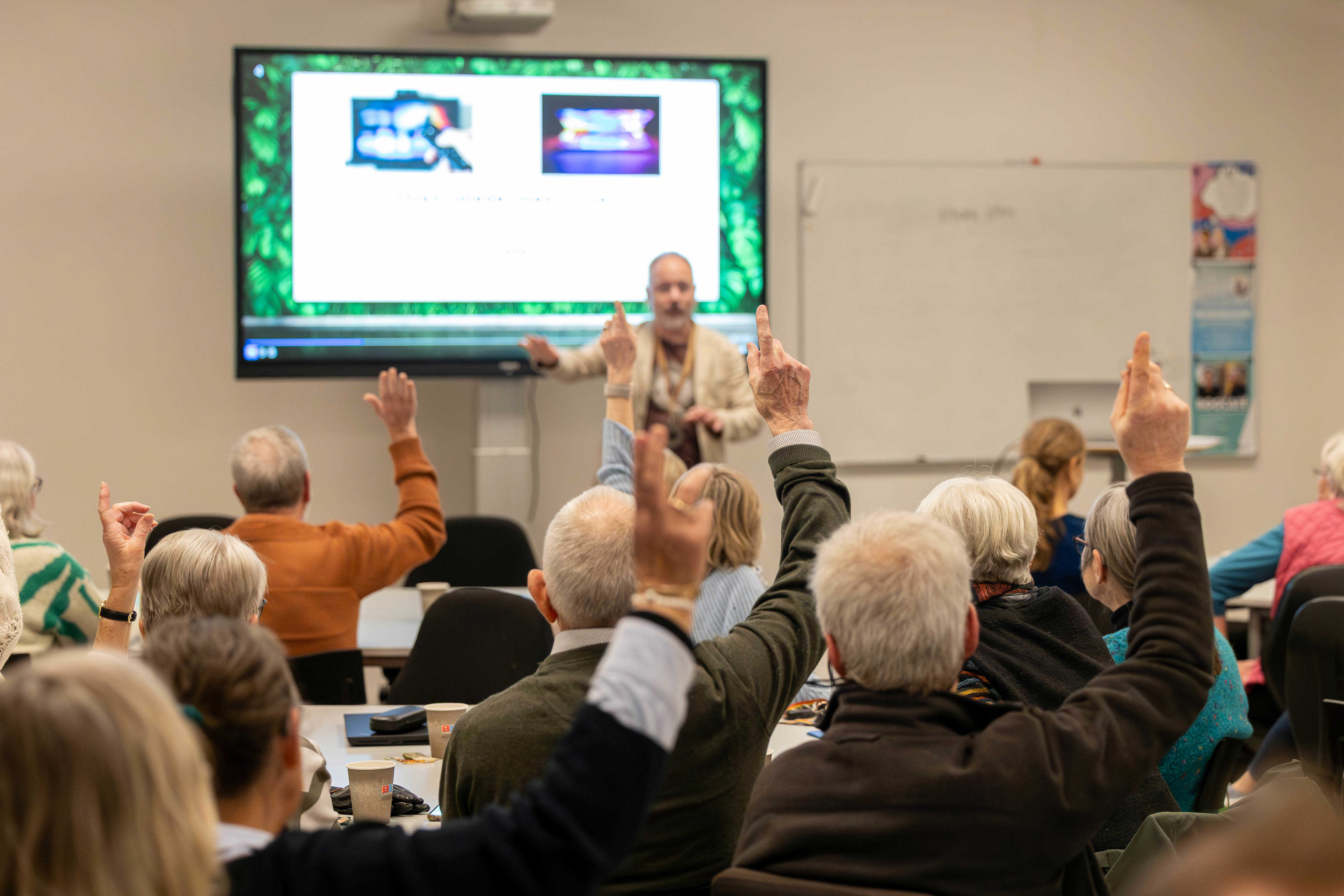 Møde. En mand står med fronten mod kamera og deltagere sidder med ryggen til og rækker hånden op